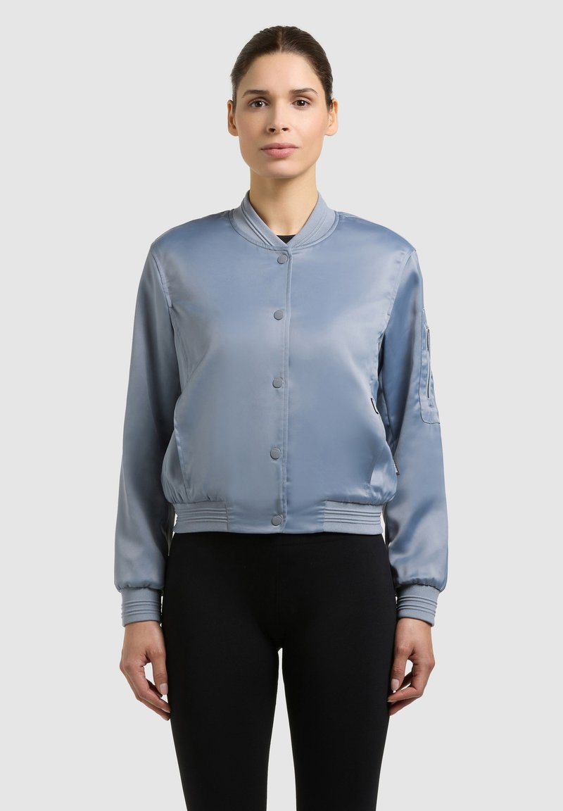 Femme portant une veste bomber en satin bleu clair avec des boutons-pression et un pantalon noir, debout devant un fond clair uni.