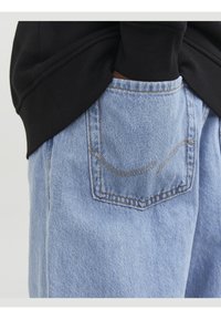 Jean en denim bleu clair avec une poche arrière présentant un détail de couture subtile et courbée, associé à un sweat-shirt noir.