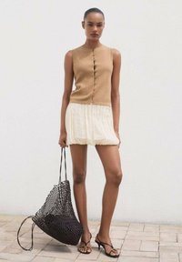 Beige bezrukávový top s předními knoflíky, kombinovaný s bílou plisovanou sukní. Modelka drží černou tkanou tašku a má na sobě černé sandály na podpatku.