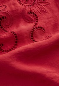 Tissu rouge avec des frivoles broderies complexes et des motifs en dentelle découpée, montrant la texture et les détails de couture fines.