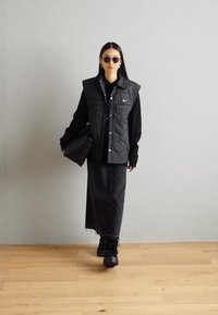 Black padded vest with two front pockets, worn over a black long-sleeve top and a dark skirt. Black platform shoes and round sunglasses complete the look.
