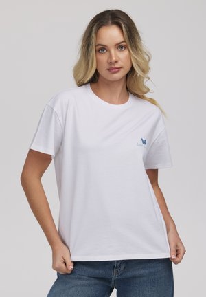 Kobieta z blond włosami do ramion, ubrana w białą koszulkę z małym niebieskim motylem i napisem na lewym biuście, w parze z niebieskimi dżinsami.