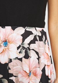 Zwarte jurk met een aansluitende top en een bloemenrok. Bevat grote roze bloemen met blauwe hartjes en groene bladeren op een donkere achtergrond.