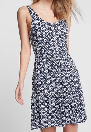 Femme portant une robe bleu marine sans manches avec un motif floral géométrique blanc et de petits boutons sur le devant.