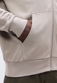 Χέρι κρυμμένο στην μπροστινή τσέπη ενός μπεζ φλις με φερμουάρ που φοριέται πάνω από σκούρο παντελόνι.