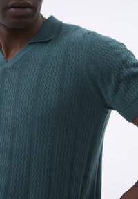 Texturovaná tyrkysová pletená polo košile s vertikálními pruhy a krátkým límcem. Materiál má prodyšný, otevřený tkaný design.