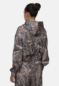 Hoodie camouflage arborant un mélange de gris foncés, de bruns et de motifs de feuilles. Conçu avec une capuche et des manches longues, offrant une coupe décontractée.