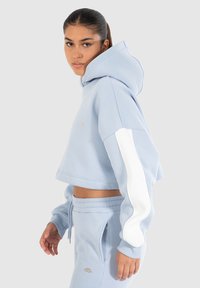 Sudadera corta de color azul claro con una gran capucha, rayas blancas en los laterales y puños acanalados. El material es suave y tiene un corte relajado.