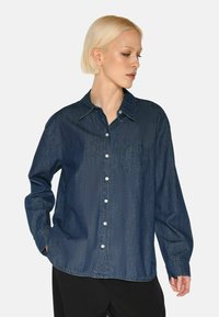 Camicia di denim blu scuro, con chiusura a bottoni, colletto a punta, maniche lunghe e una tasca sul petto. Texture liscia.