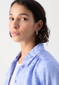 Lys blå bluse med blomstermønster, knappe-lukking og strukturert krage. Modellen har på seg hoop øredobber og har bølgete, skulderlangt hår.