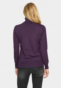 Femme aux cheveux blonds longs portant un pull à col roulé violet et un jean noir, debout de dos contre un fond blanc uni.