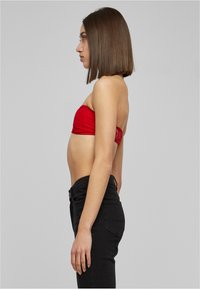 Sarkans strapless tops ar teksturētu audumu, apvienots ar augstām melnām džinsam. Sānu profils parāda pieguļošu siluetu un nav redzamu rakstu.