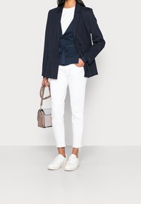 Navy blazer over a dark blue wrap top, paired with white tailored pants and white sneakers. Holding a beige and gray handbag.