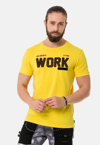 Gelbes Baumwoll-T-Shirt mit großem schwarzem "WORK"-Druck, Rundhalsausschnitt und kurzen Ärmeln, kombiniert mit gemusterten grauen und schwarzen Shorts.