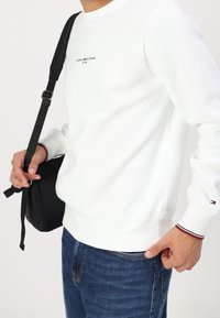Biała bluza z okrągłym dekoltem i subtelnym logo. Czarna torba typu crossbody z regulowanym paskiem. Niebieskie dżinsy o luźnym kroju.