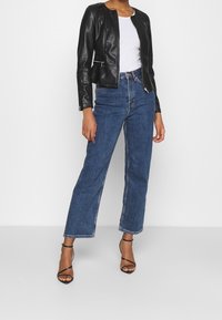 Giacca nera in ecopelle con collo rotondo, abbinata a jeans di jeans blu a gamba larga e vita alta e sandali neri con cinturini.