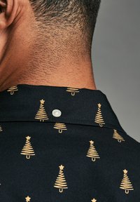 Chemise noire avec un motif d'arbre de Noël doré, dotée d'un bouton blanc au col. Tissu texturé avec une coupe ajustée.