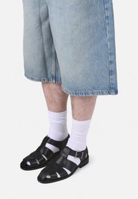 Sandales en cuir noir avec un design tissé ouvert, détail de boucle en métal, portées avec des chaussettes blanches et un short en denim bleu clair.