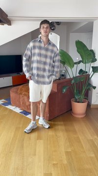 Camicia a quadri chiari con maniche lunghe, pantaloni corti bianchi e sneakers grigie. Presenta un divano in velluto a coste marrone e piante verdi in una stanza ben illuminata.