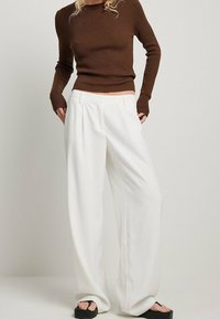 Modellen står i avslappnade vita byxor och en figurnära brun långärmad topp, och visar upp en modern, avslappnad outfit med svarta sandaler.