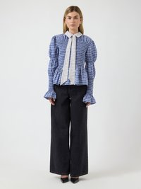 Γυναίκα που φοράει μπλε καρό μπλούζα με φουσκωτά μανίκια, λευκή καρό γραβάτα και μαύρο τζιν με φαρδιά πατζάκια, στέκεται μπροστά σε λευκό φόντο.