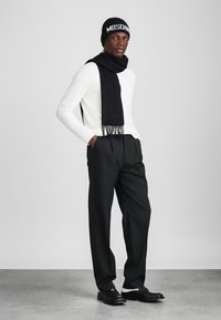 Melna adīta cepure ar "MOSCHINO" logotipu, melna šalle ar frīzi, baltas garām piedurknēm krekls, melnas bikses un melnas kurpes. Vienkāršs, moderns dizains.