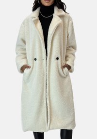 Roomkleurige, oversized jas gemaakt van zachte, gestructureerde stof. Heeft een kraag, twee voorszakken en dubbele zwarte knopen.