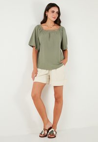 Olive green blouse with a square neckline and flutter sleeves, featuring decorative lace trim. Paired with beige shorts and embellished sandals.