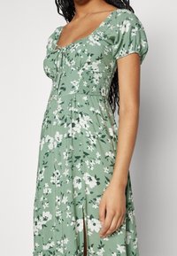 Zielona sukienka w kwiaty z krótkimi bufkami, kwadratowym dekoltem i marszczonym gorsetem. Posiada rozcięcie sięgające uda oraz teksturowaną tkaninę.