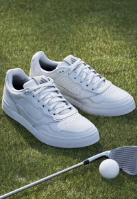 White leather sneakers with perforated detailing and round toes, displayed on grass alongside a golf club and ball.