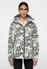 Kobieca wodoodporna kurtka, lekka, biała z czarnym abstrakcyjnym nadrukiem w panterkę, z pełnym zamkiem, kapturem, elastycznymi mankietami i dolnym brzegiem. Gładka tekstura.
