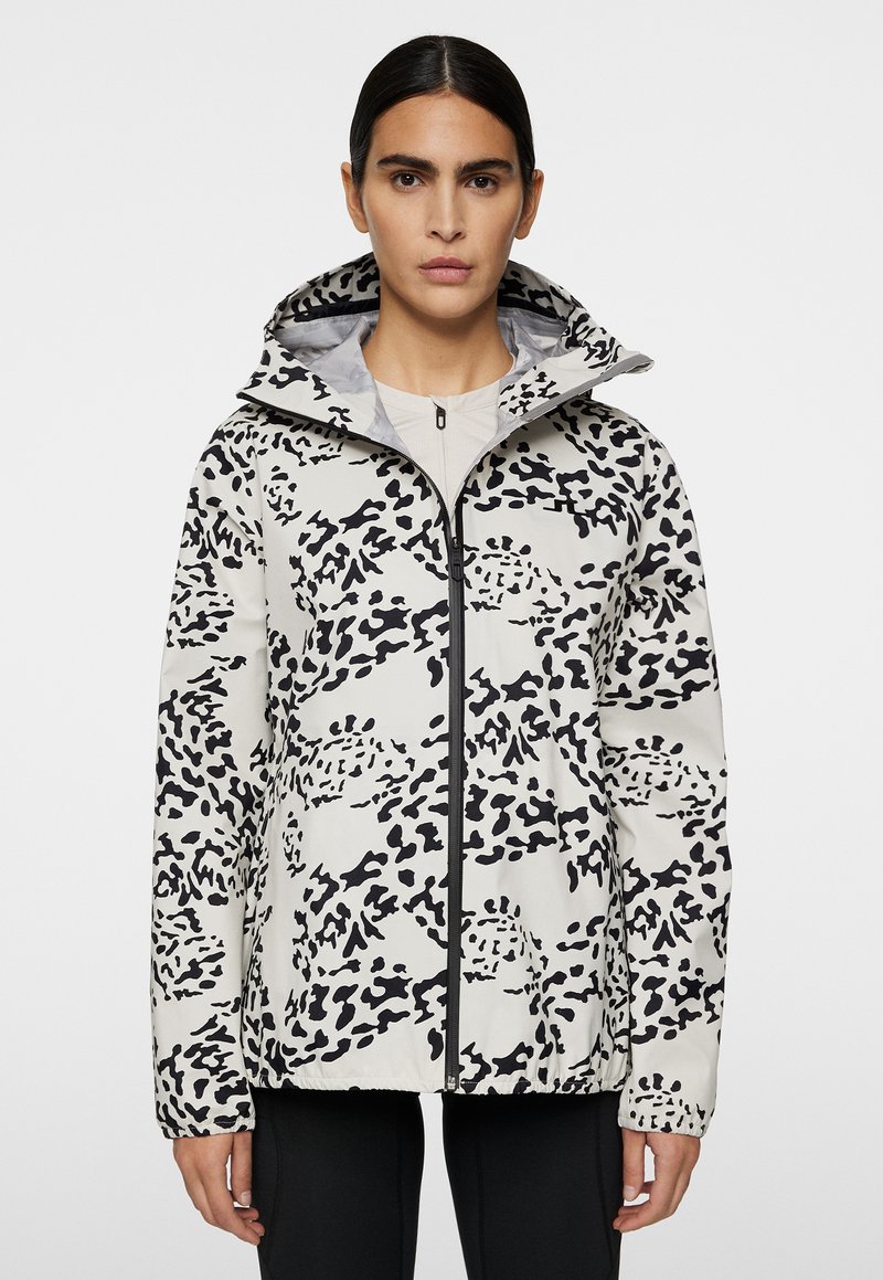Kobieca wodoodporna kurtka, lekka, biała z czarnym abstrakcyjnym nadrukiem w panterkę, z pełnym zamkiem, kapturem, elastycznymi mankietami i dolnym brzegiem. Gładka tekstura.