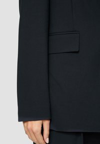 Black tailored blazer with a structured shape, textured fabric, and a subtle sheen. Features a single chest pocket and streamlined cuffs.