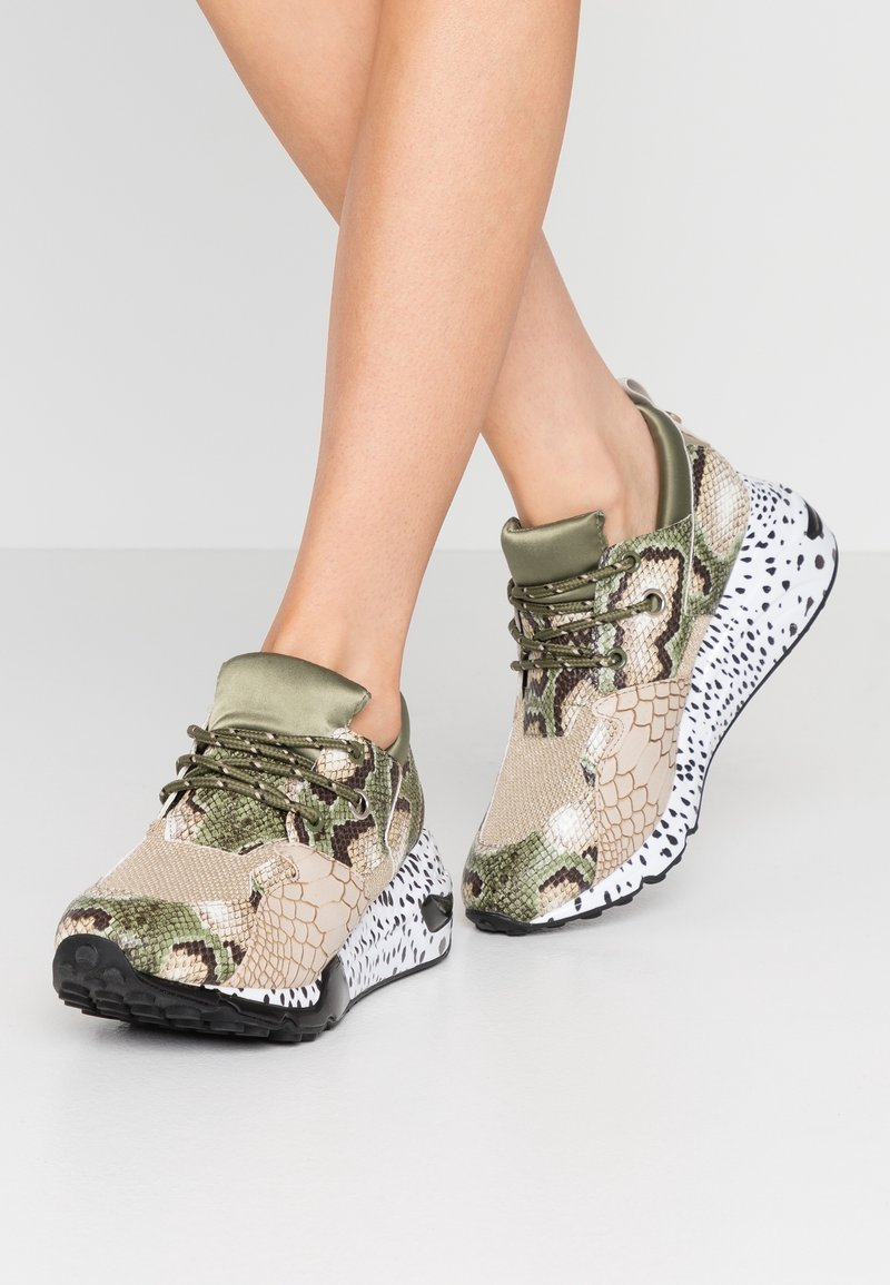 Jambes portant des baskets à plateforme imprimé serpent vert et beige avec des semelles tachetées noires et blanches, debout sur un sol gris clair.