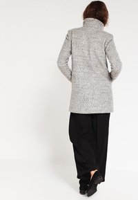 Cappotto lungo grigio texturizzato con colletto alto, vestibilità dritta e maniche lunghe. Abbinato a pantaloni neri larghi e scarpe scure.