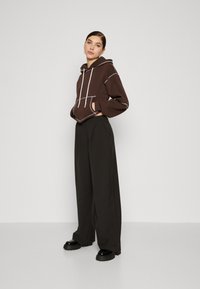 Brun cropped hettegenser med lys grå søm og frontlomme, kombinert med svarte vide bukser og kraftige svarte sko. Jevn stofftekstur.