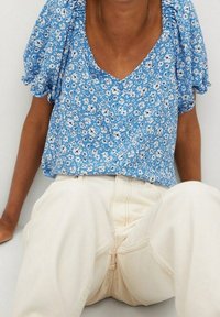 Blouse bleue à manches courtes avec un motif floral blanc et un décolleté en V, associée à un jean crème taille haute. Tissu texturé avec des manches volantes.