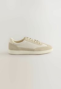 Zapatilla deportiva beige con parte superior de malla texturizada y acentos de ante suave. Cuenta con una punta redondeada y una suela blanca contrastante.