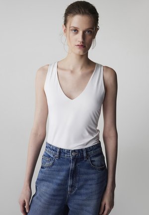 Donna con pelle chiara e capelli castani raccolti, che indossa un top bianco senza maniche a scollo a V e jeans di denim blu a vita alta su uno sfondo semplice.