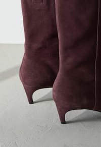 Burgundské semišové ankle boty s špicatou špičkou, úzkym stiletto podpätkom a minimálnymi detailmi šitia. Hladká textúra s matným povrchom.