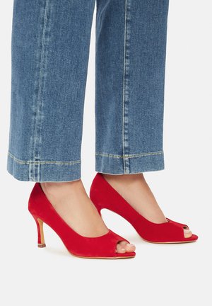 Rote offene Sandaletten aus glattem Wildleder mit einem spitzen Absatz, ergänzt durch eine goldakzentuierte Absatzbasis. Kombiniert mit blauen Jeanshosen.