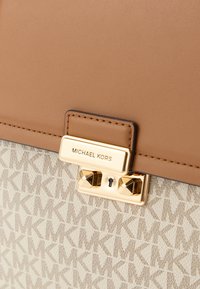 Brązowy element torebki ze skóry, z beżową, fakturowaną podstawą z wzorem "MK", złotym okuciem i detalem zamka na zatrzasku.