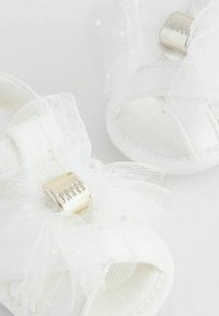 White sandals featuring a large tulle bow with polka dots and a metallic accent. The design includes a soft, rounded shape and smooth texture.