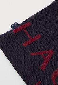 Fular întunecat de culoare bleumarin, cu text roșu îndrăzneț, textură tricotată și o etichetă mică de branding cu inscripția "HACKETT" pe o margine.