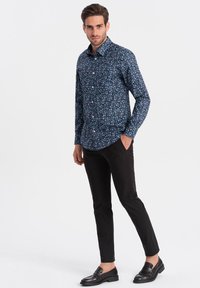 Camicia button-up con sfondo blu navy, caratterizzata da un motivo floreale in vari colori. Indossata con pantaloni neri e mocassini in pelle.