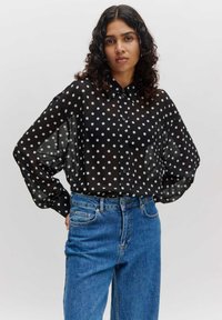 Zwarte sheer blouse met witte polka dots, knopen aan de voorkant en een losse pasvorm. Gecombineerd met hoogzitende blauwe denim jeans.