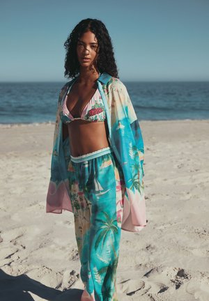 Junge Frau mit lockigem Haar, die einen farbenfrohen Bikini mit tropischem Muster und passende lockere Hosen sowie ein Überwurf trägt, steht an einem Sandstrand am Meer.