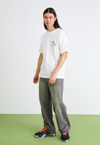 Un jeune homme aux longs cheveux noirs se tient sur un sol vert, portant un t-shirt blanc avec trois petits graphiques, un pantalon gris et des baskets noires avec des lacets orange.