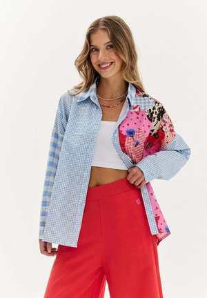 Koszula w wielu kolorach, z niebieską kratą gingham i wzorami kwiatowymi, o luźnym kroju. Połączona z czerwonymi spodniami, które mają lekką strukturę i wdzięk.