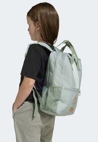 Zeleni ruksak s teksturiranim završetkom, s dvije mrežaste bočne džepove i tri bijele pruge. Model nosi crnu majicu i sive hlače.